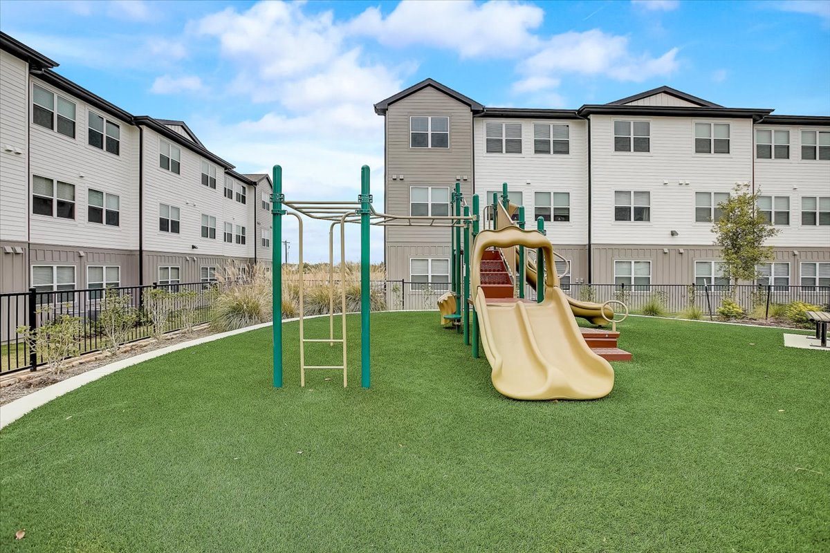 Community playground with slides and climbing features on soft turf, set between apartment buildings.